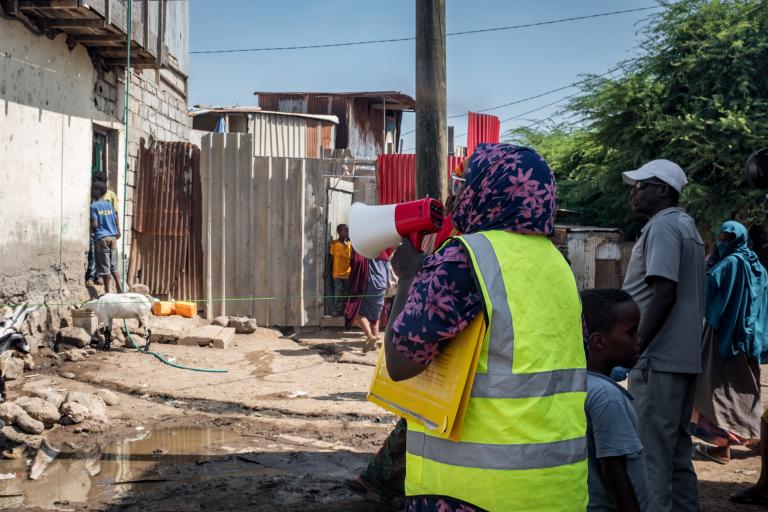 A person in a yellow vest holds a megaphone and documents while addressing people in an informal settlement area with scattered debris and makeshift structures.
