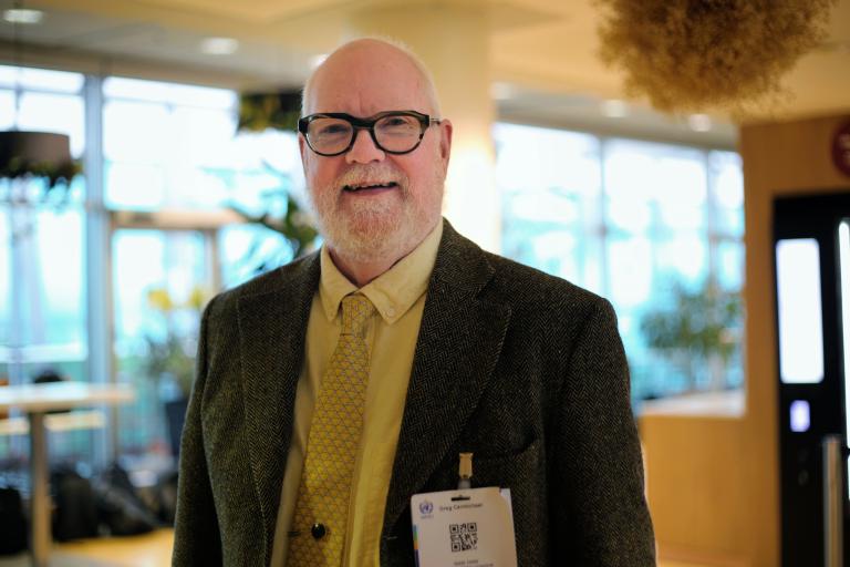 Bald man with glasses, beard, and mustache wears a suit, yellow shirt, and tie, standing indoors with a conference name badge clipped to his jacket.