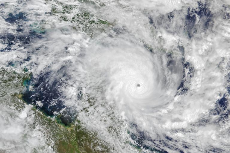 Satellite view of a large hurricane with a well-defined eye over the ocean, surrounded by thick clouds and spiral bands extending toward nearby landmasses.