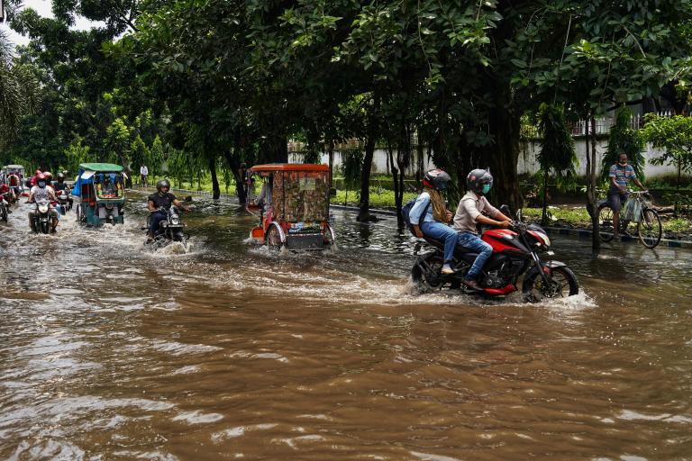 Motorcycles, rickshaws, and bicycles travel through a heavily flooded street lined with trees during daytime.