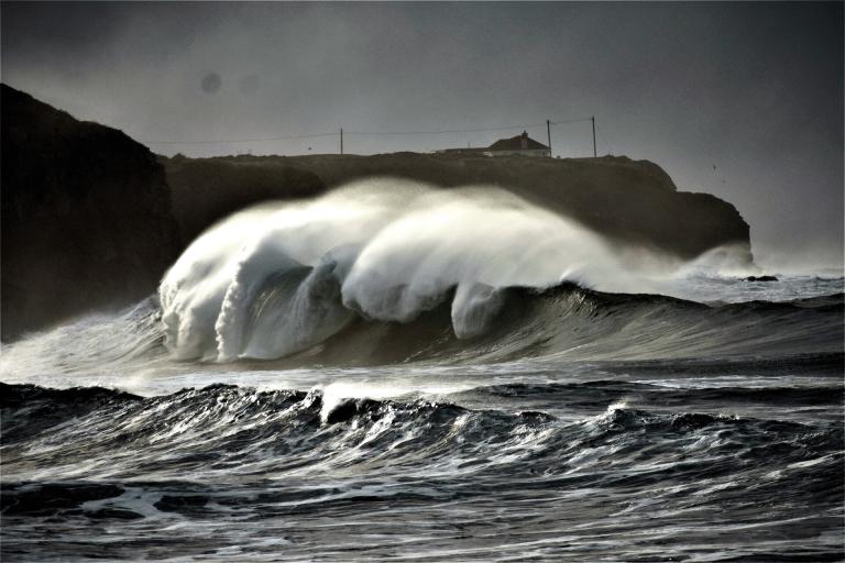 Large ocean waves crash near a rocky coast under a cloudy sky, with a silhouette of a cliff and buildings in the background.
