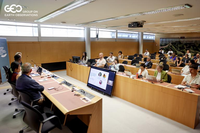 People attend a conference in a modern meeting room, with speakers at a front panel and a large screen displaying a presentation; GEO Group on Earth Observations logo is visible.
