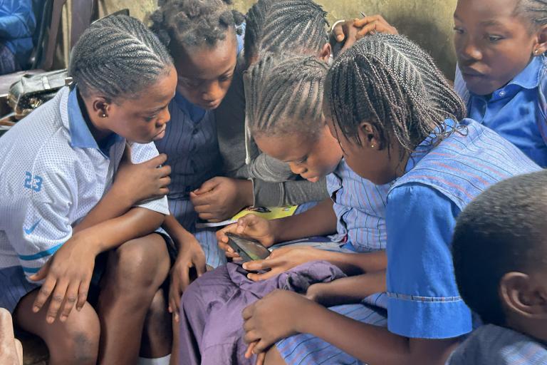 A group of schoolgirls in uniforms sit closely together, looking intently at a smartphone, inside a classroom.