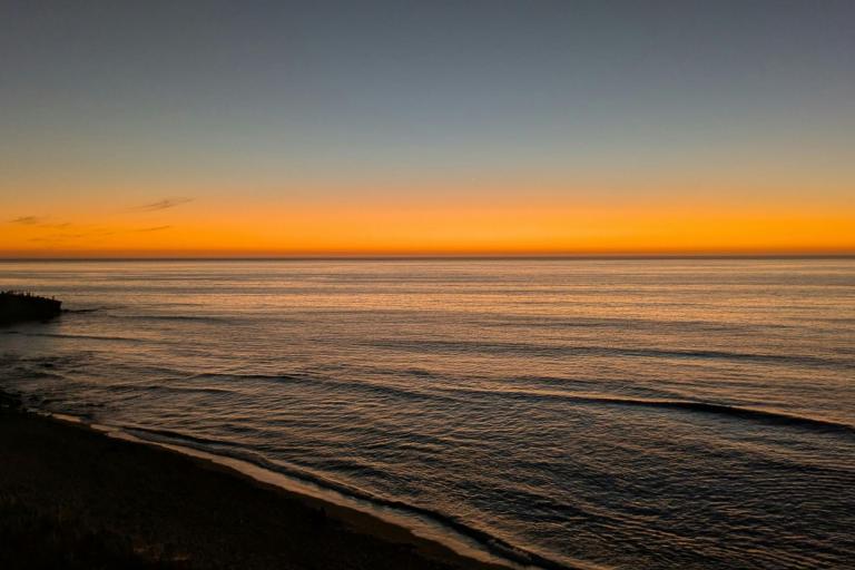 Calm ocean waves under a clear sky at sunset, with an orange glow on the horizon and silhouettes of land on the left side.