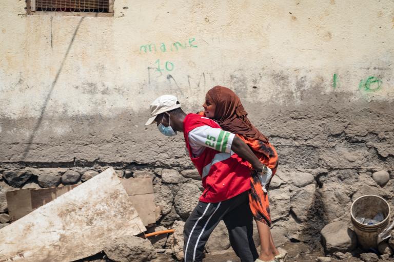 A person in a red vest and face mask carries another person on their back along a rough, debris-strewn street next to a worn wall.
