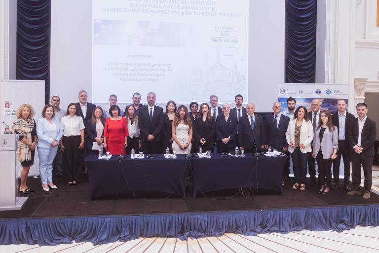 A group of about 25 people in business attire pose for a photo in a conference room in front of a screen displaying meeting details.