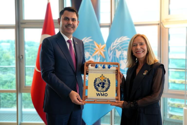 Two people stand indoors holding a framed WMO emblem, with the Turkish and WMO flags visible in the background.