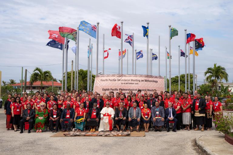 A large group of people pose for a photo in front of international flags and a banner for the Pacific Meteorological meeting, with palm trees and a clear sky in the background.