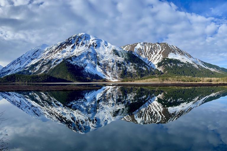 Snow-capped mountains are reflected in a calm lake under a blue sky with scattered clouds.