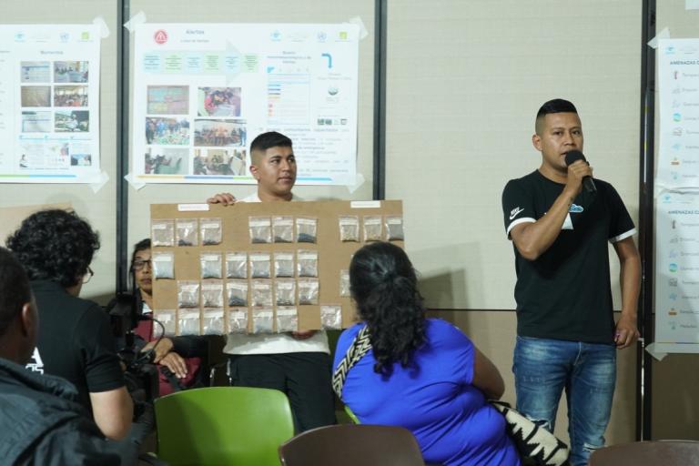 A man speaks into a microphone while another holds a display board with many small bags. Several people are seated, and posters with images and charts are on the wall behind them.