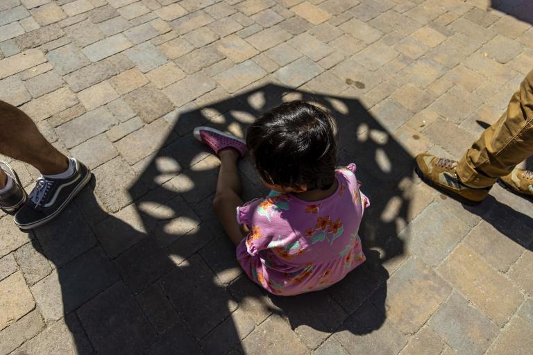 A child in a pink floral dress sits on a stone pavement, partially shaded by an umbrella, with three adults’ legs visible nearby.