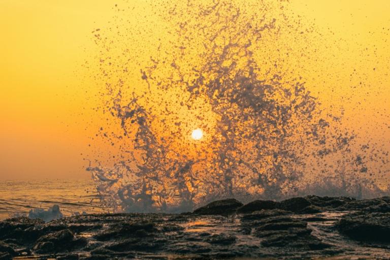 Ocean waves splash against rocks at sunset, with the sun visible through water droplets and an orange sky in the background.