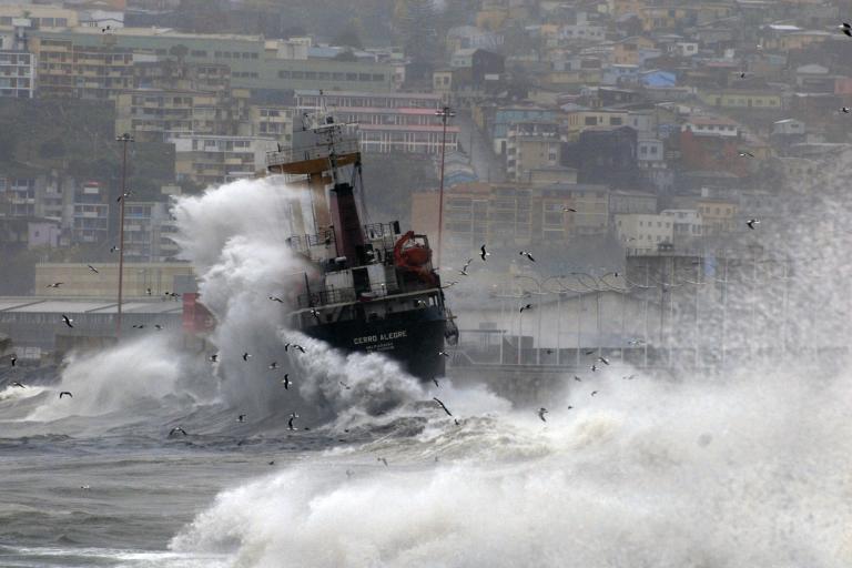 A ship being struck by large waves in a harbor, with a hillside city in the background.