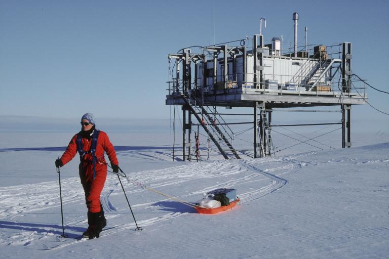 A person in a red suit cross-country skis across snow, pulling a loaded orange sled toward a raised research building.