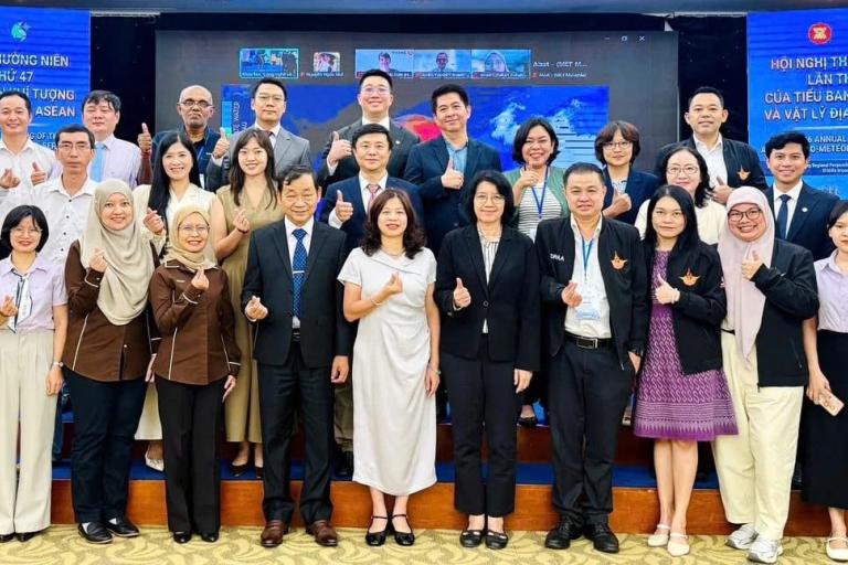 A large group of people in business attire posing for a photo on a stage in front of a digital screen displaying text about an ASEAN meeting.