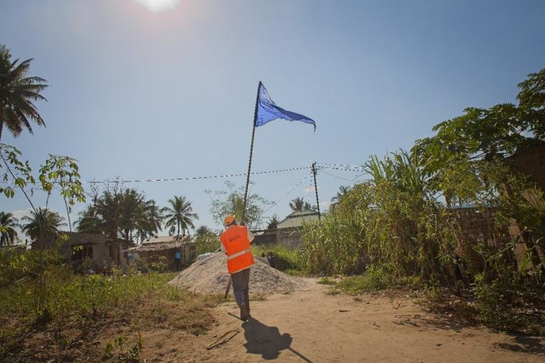 A person in an orange vest carries a tall pole with a blue flag on a dirt path, with trees and houses in the background under a clear sky.