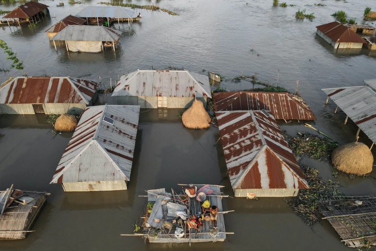 Aerial view of houses partially submerged in floodwater, with people on a makeshift raft in the center.