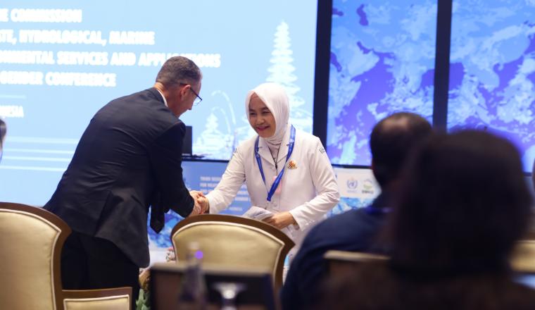 Two professionals shaking hands at a conference with a presentation screen in the background.