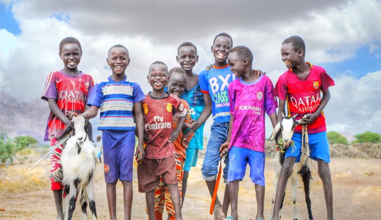 A group of seven children stands outdoors on a dirt path, smiling at the camera. Two of the children are holding goats. The sky is partly cloudy in the background.