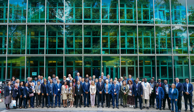 A large group of individuals in formal attire stands in several rows in front of a modern glass building, posing for a group photo.