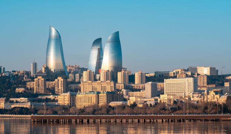Skyline of a city featuring three flame-shaped glass towers and surrounding buildings reflected in a calm body of water.