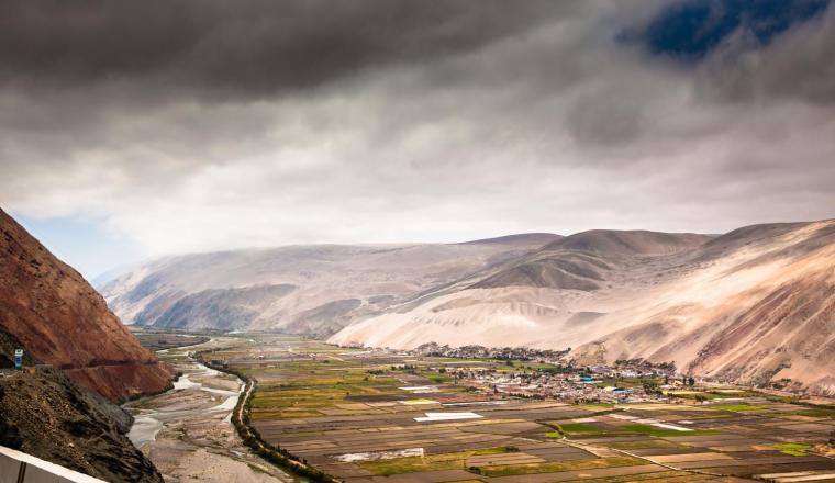 A scenic view of a river winding through agricultural fields and a small settlement, surrounded by rolling hills under a cloudy sky.