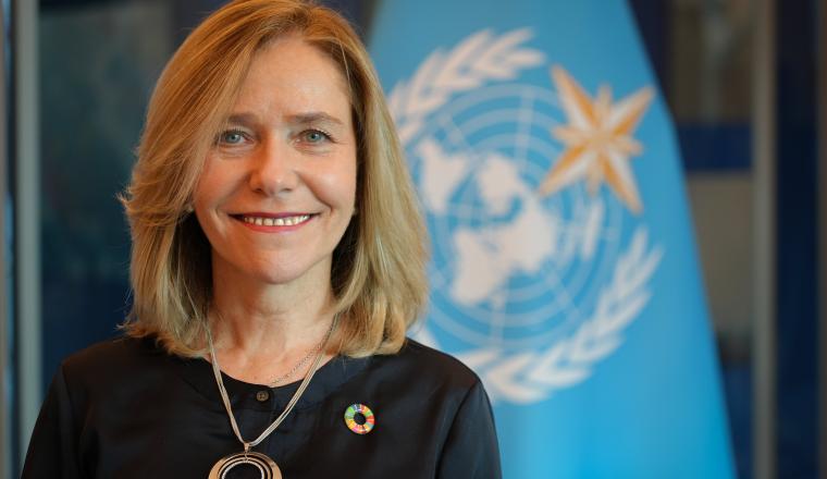 Smiling woman with shoulder-length blonde hair wearing a black top and necklace, standing in front of a blue United Nations flag with a circular pin.