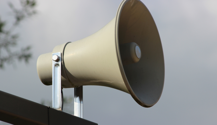 A close-up of a gray outdoor loudspeaker mounted on a metal bracket against a cloudy sky background.