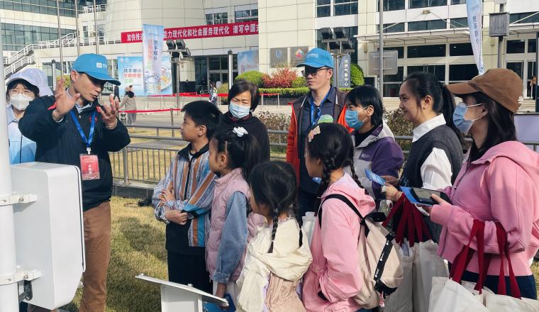 A group of adults and children, some wearing masks, listen attentively to a person speaking outside a modern building. The scene suggests an educational or guided tour.