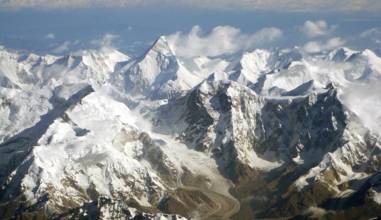 Aerial view of snow-covered mountain peaks with a glacier winding through the valley below under a partly cloudy sky.