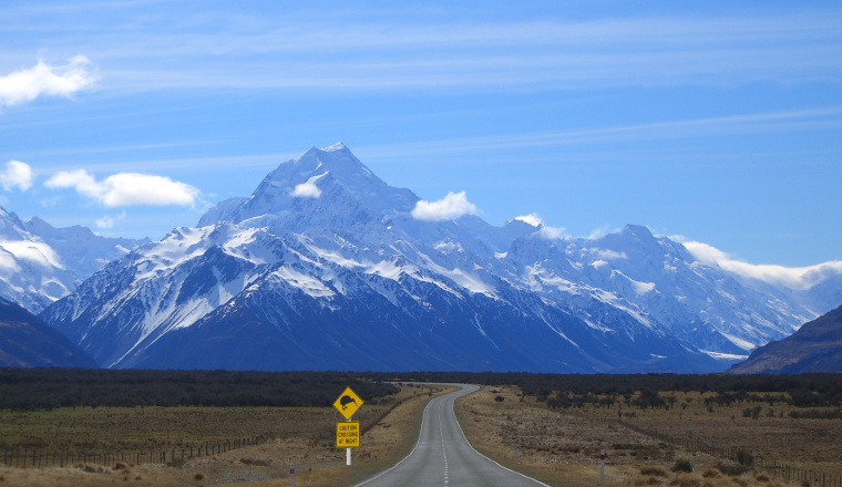 A straight road leads toward snow-covered mountains under a blue sky, with a yellow road sign on the left side of the road in the foreground.