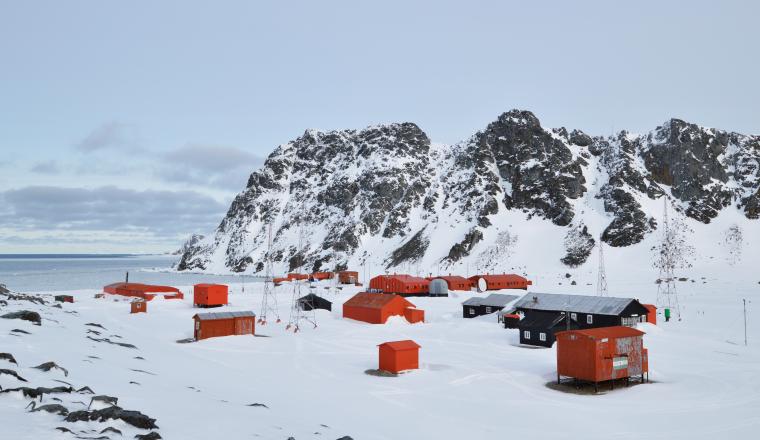 A cluster of red and black buildings sits on snow-covered ground near rocky, snow-capped mountains and the sea under a cloudy sky.