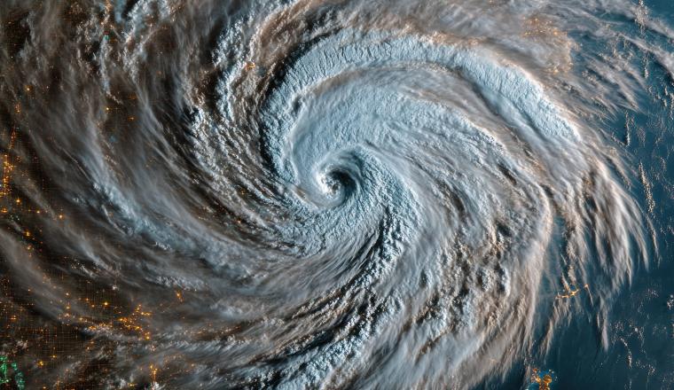 Satellite view of a large hurricane swirling over land and ocean, with city lights visible beneath the clouds on the left side of the image.
