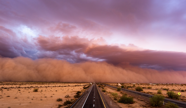 A large dust storm approaches a deserted highway in a desert landscape under a dramatic, cloudy sky at sunset.