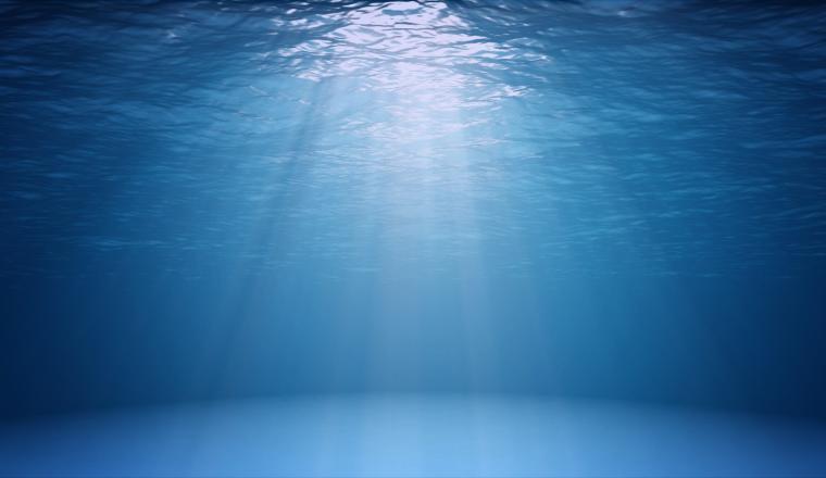 Sunlight filters through clear blue ocean water, casting rays onto the calm, sandy seafloor below.