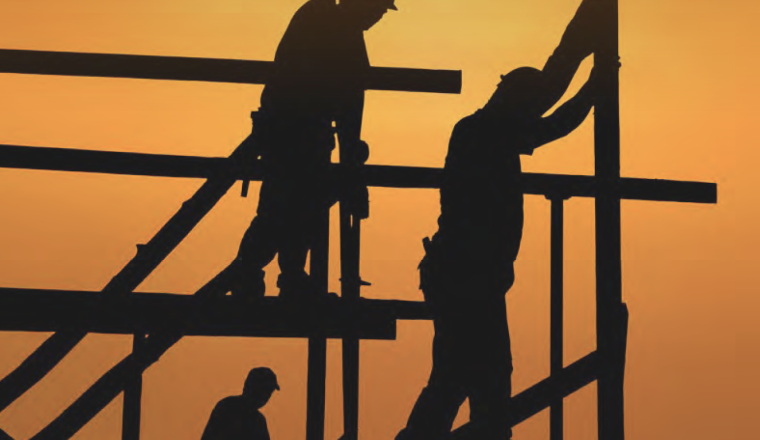 Three construction workers are silhouetted against an orange sunset sky as they work on a building framework.