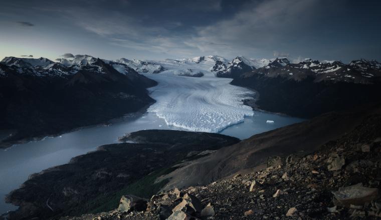 A large glacier flows between mountain ranges, ending in a lake under a cloudy sky, with rocky terrain in the foreground.