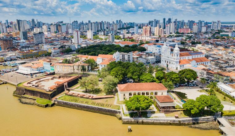 Aerial view of a historic riverside district with colonial buildings, a fort, and modern city skyline in the background under a partly cloudy sky.