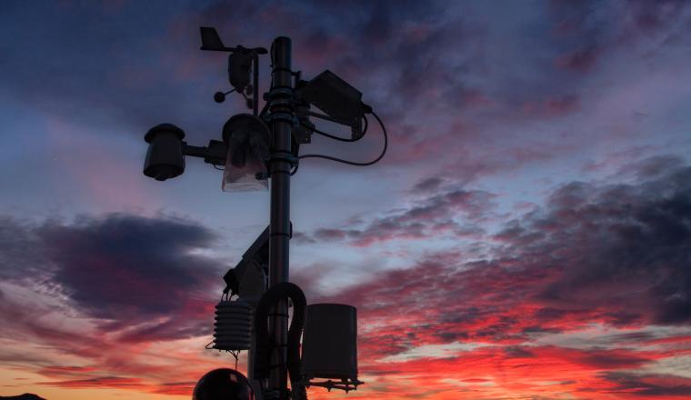 A weather station with various sensors stands silhouetted against a dramatic sunset sky with red, orange, and blue clouds.