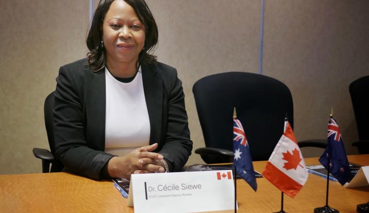 A woman in a black blazer sits at a conference table with small Canadian, Australian, and New Zealand flags and a nameplate reading "Dr. Cécile Siewe.
