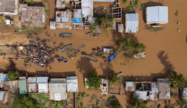 Aerial view of a flooded village with submerged houses, boats, and a large group of people gathered on a dry patch of ground.