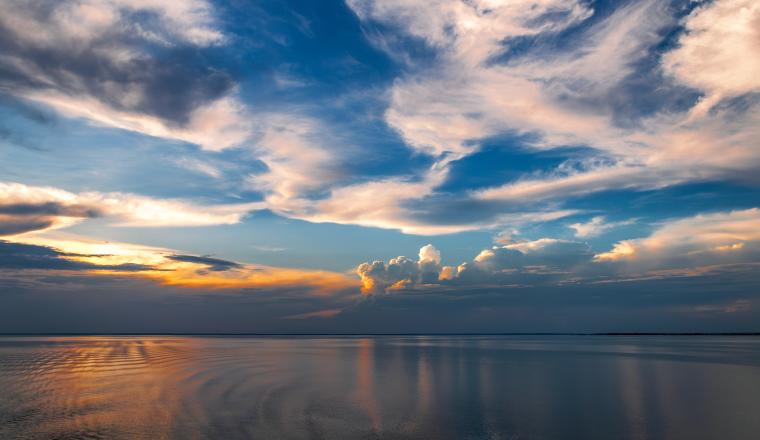Calm water reflects a colorful sky with scattered clouds at sunset, creating a peaceful and open landscape.