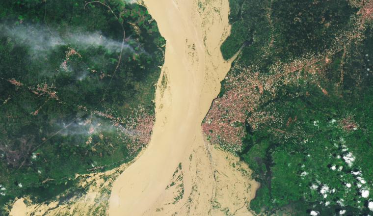 A wide, muddy river separates two densely built urban areas, surrounded by green vegetation and patches of cloud cover, as seen from above.
