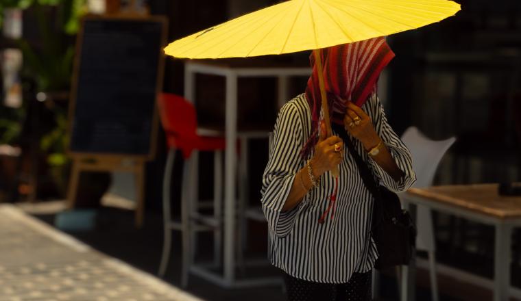 Person holding a large yellow umbrella, face covered by a striped scarf, standing on a sunlit sidewalk near outdoor seating and a blackboard sign.