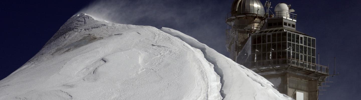 Snowy mountain peak with wind-blown snow and an observatory building featuring a dome and antennas under a clear blue sky.