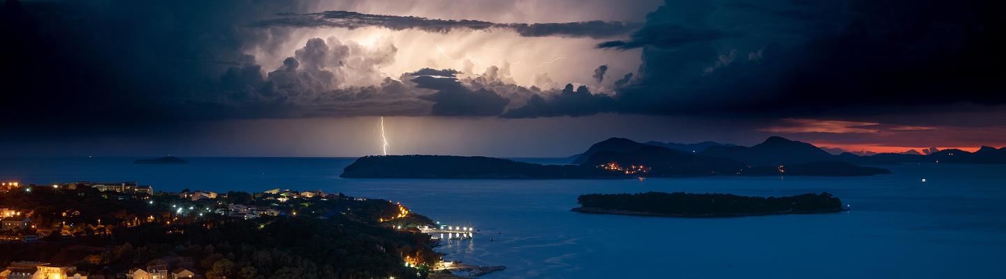 Dramatic lightning illuminates storm clouds above a coastal town at night, with islands and a cruise ship visible on the calm sea.
