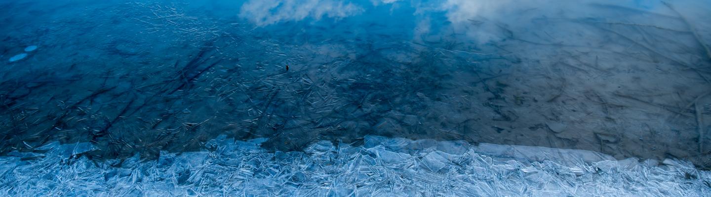 A partially frozen lake with ice crystals forms in the foreground, reflecting blue sky and white clouds.