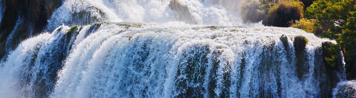 Multilevel waterfall cascading over rocky terrain surrounded by lush greenery under a clear sky.
