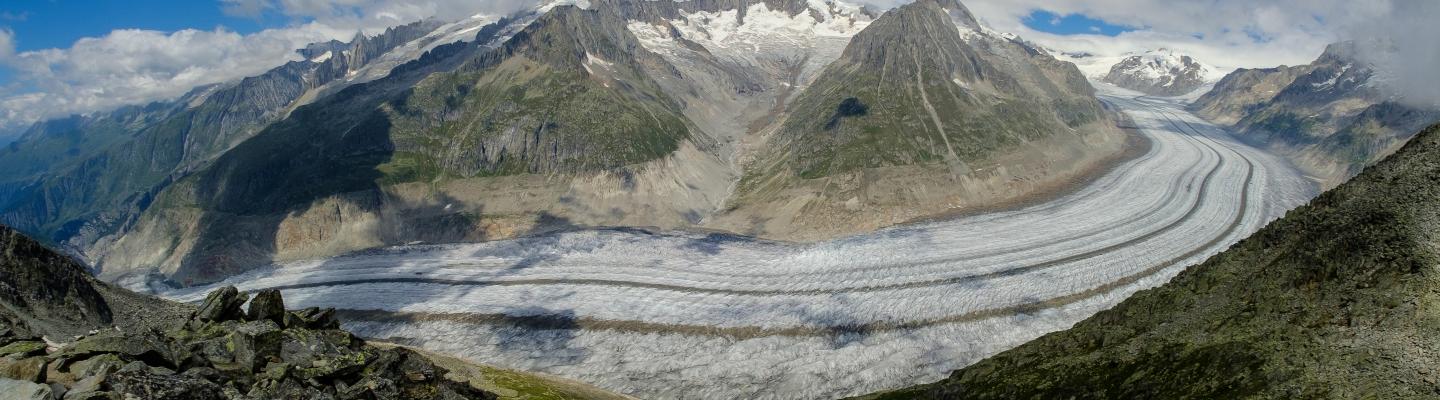 Panoramic view of a glacier winding between rugged mountains under a blue sky with scattered clouds. Rocky foreground leads to the expansive ice flow, surrounded by green slopes and rocky peaks.