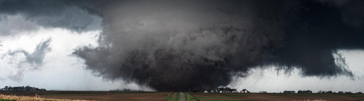 A large, dark tornado approaches a rural road, flanked by open fields under an ominous sky.